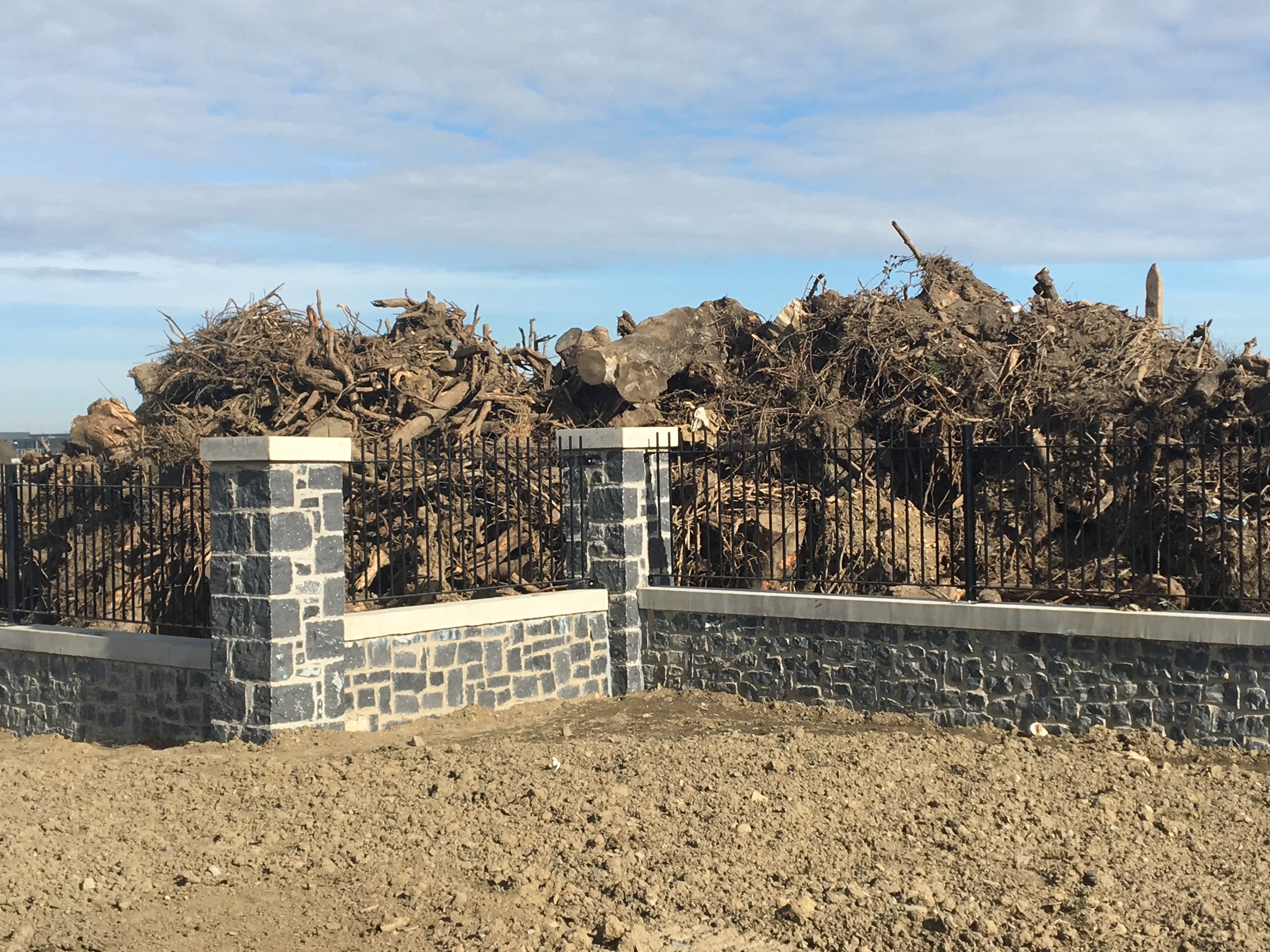 A pile of tree roots and pieces sits behind a prefab stone and metal fence.