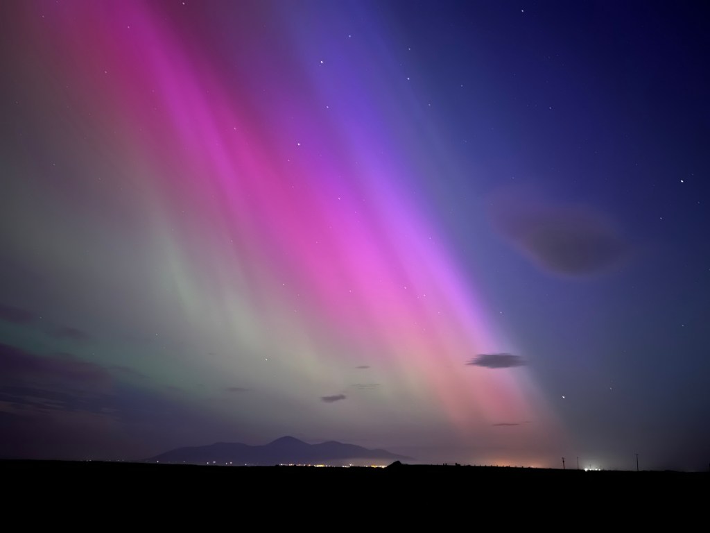 A night sky, with the Mourne Mountains on the horizon, silhouetted by an auroral curtain.