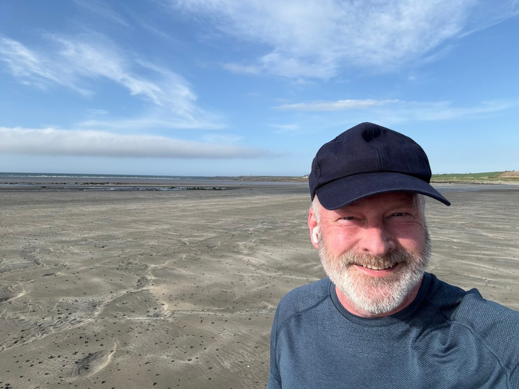 A selfie of the author, a smiling man with a short grey beard and a baseball cap. Behind him is a sandy beach stretching to the horizon, where it meets a blue, cloud-speckled sky.